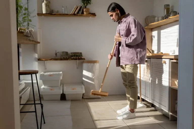 Man with long hair in long sleeve shirt clean kitchen area sweeping